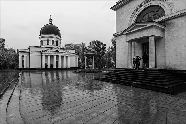 O reflectare a unei clădiri pe o curte umedă, din cărămidă, într-o fotografie alb-negru.