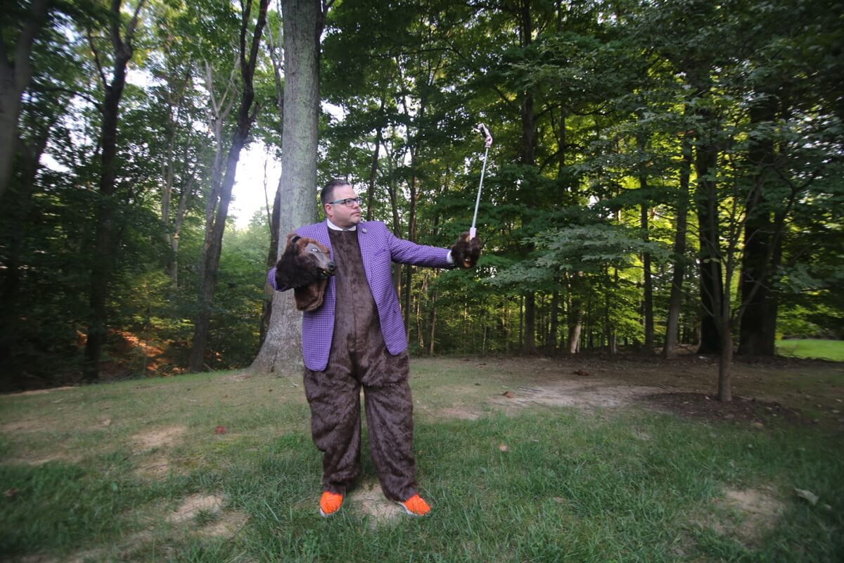 Jay Baer dalam setelan beruang berdiri di atas rumput dengan hutan di latar belakang.