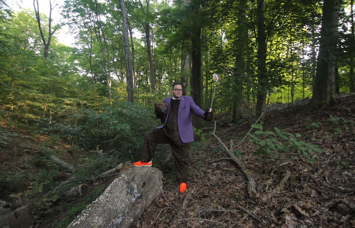Jay Baer mengenakan setelan beruangnya berdiri di hutan lebat dengan kakinya di atas kayu dan tongkat selfie mengambil fotonya