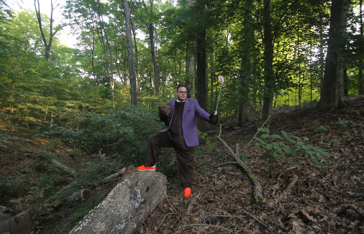 Jay Baer indossa il suo costume da orso in piedi in una fitta foresta con il piede su un tronco e un bastone per selfie che scatta la sua foto