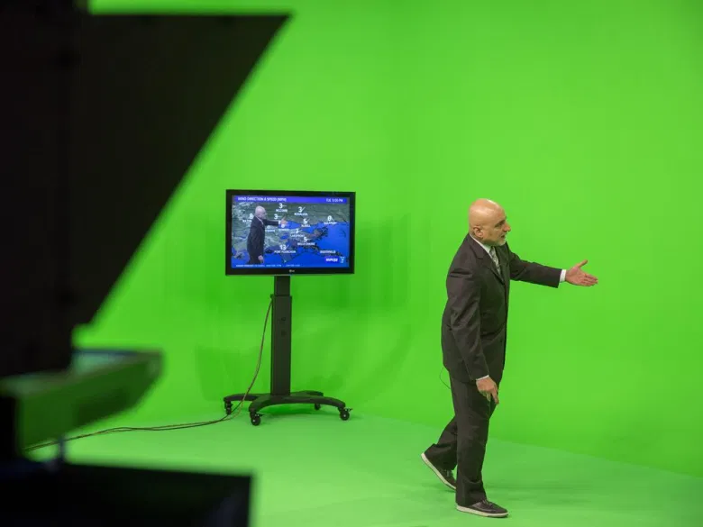 Un prezentator meteo care stă în fața unui ecran verde. Un monitor TV arată cum arată el suprapus peste harta vremii.