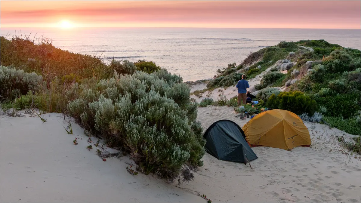 Person am Strand von Margaret River in Australien, die neben zwei Zelten steht und den Sonnenuntergang betrachtet.