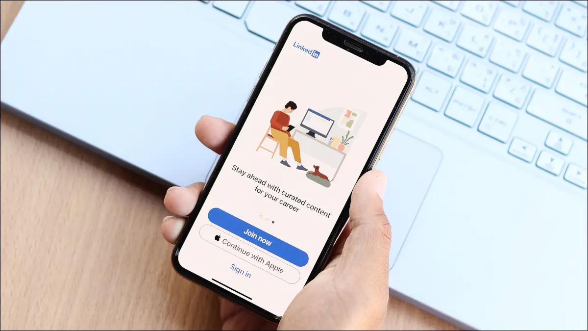 Mão de uma pessoa segurando um smartphone sobre um teclado de laptop, com o aplicativo LinkedIn em exibição.