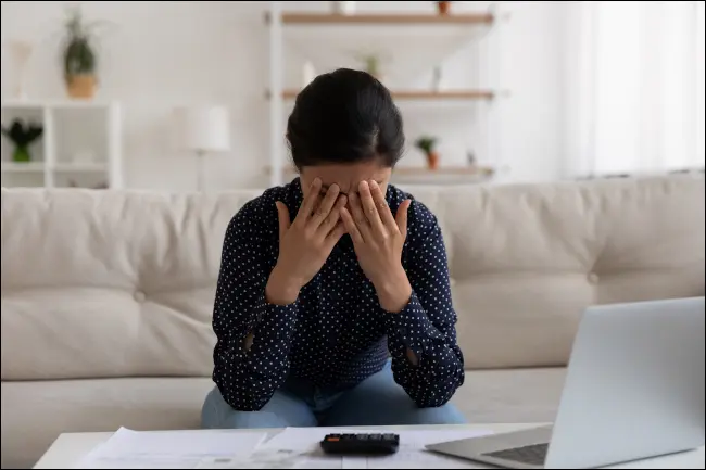 電卓とラップトップを前に置いて、額に手を押し当てて緊張している女性。