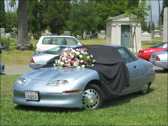 Fotografie cu mai multe mașini EV1 parcate într-un cimitir, cu flori și o draperie care acoperă una dintre mașini